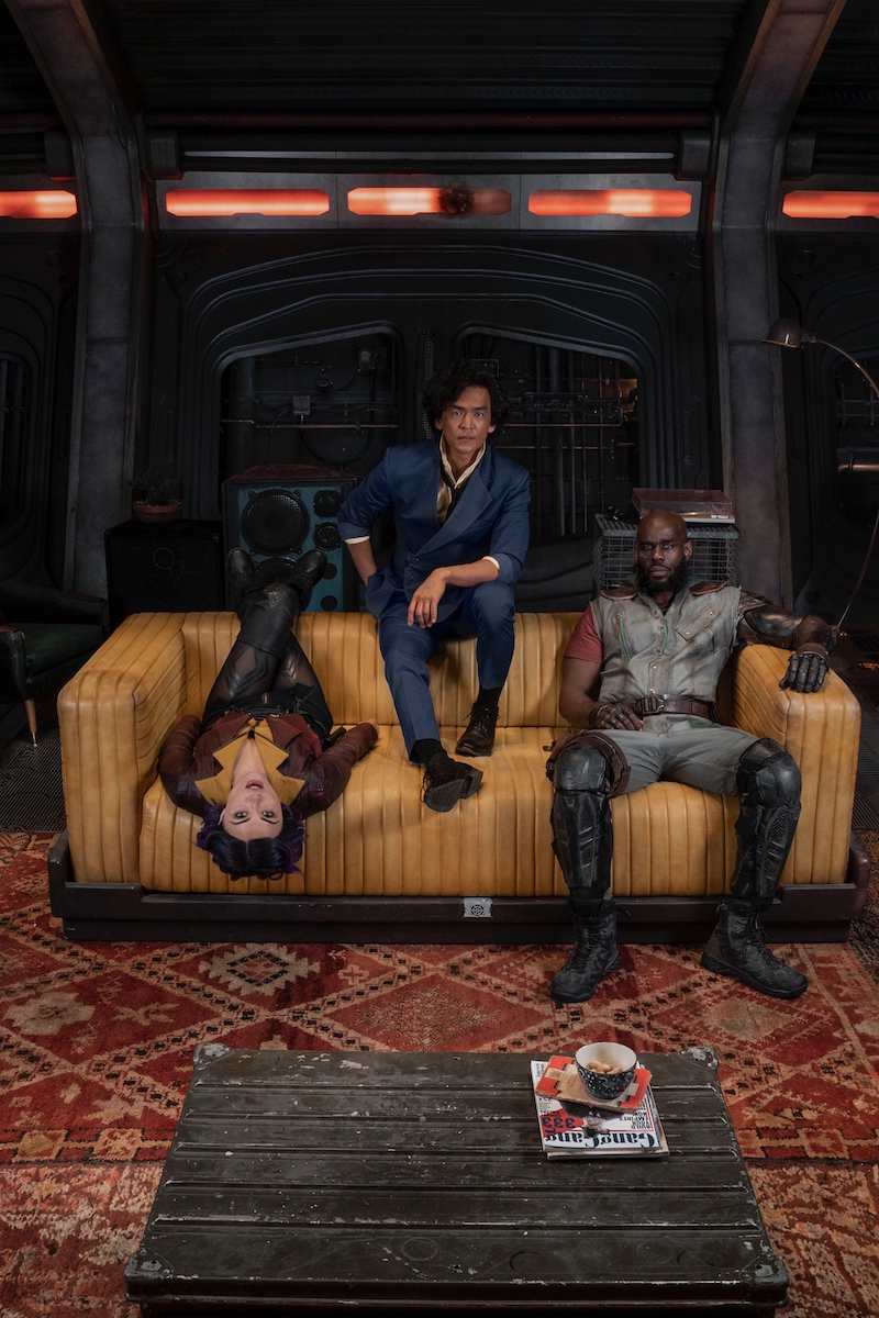 A promotional poster for Netflix's upcoming live-action Cowboy Bebop series, featuring the main cast in full costume and make-up hanging out on a couch in the hull of the Bebop space trawler.