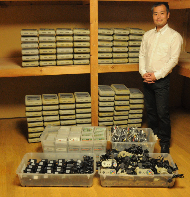 A promotional photo featuring a member of the Japan Retro Game Association posing with the 100 Super Famicom video game consoles (and their various cords, cables, controllers, and software cartridges) that the group is giving away to families impacted by the COVID-19 pandemic.