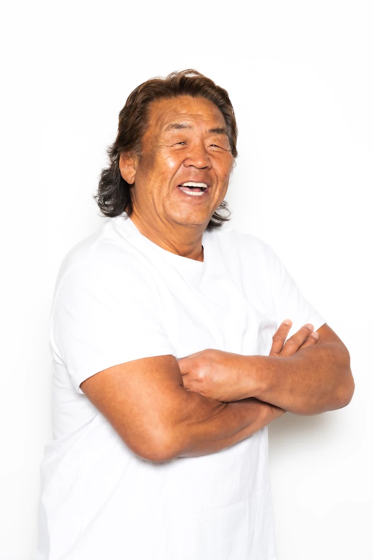 A promotional photo of retired pro wrestler Riki Choshu. Choshu smiles warmly and stands with his arms crossed while wearing a white T-shirt.