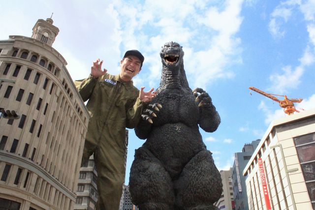 Ein Werbefoto vom Set der "Godzilla vs.  Hedorah" Kurzfilm, in dem der Regisseur Kazuhiro Nakagawa eine furchterregende Monsterpose einnimmt, während er lacht und neben dem Godzilla-Anzug aus dem Film Godzilla: Final Wars von 2004 steht.