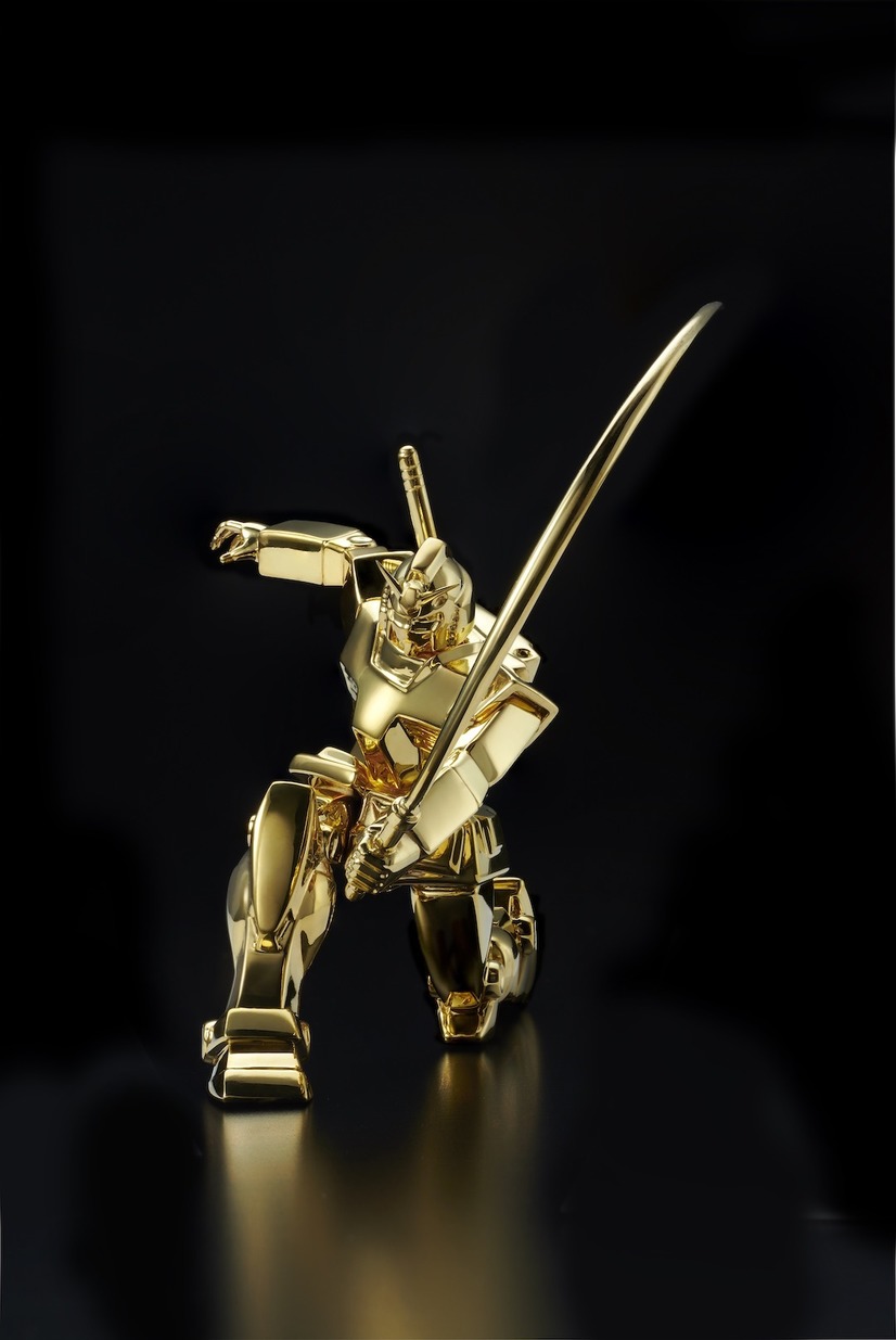 A promotional image of the The U-Works Mobile Suit Gundam Solid Gold Statue RX-78-2 Gundam (Beam Rifle Ver.), featuring the solid gold mecha crouched in a martial arts pose indicating its pilot has just cut clean through a foe.