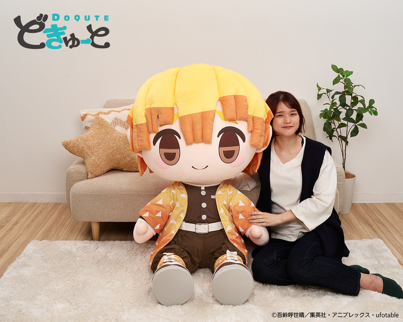 A promotional image for the DoQute 2XL Plush Toy of Zenitsu Agatsuma from Demon Slayer: Kimetsu no Yaiba by Taito Corporation. The image depicts the jumbo-sized plush toy next to a Japanese model seated in front of a sofa for a size comparison.