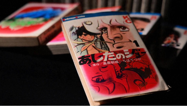 A promotional image from the upcoming Another Stories: Unmei no Bunkiten - Ashita no Joe Jidai to Ikita Hero documentary featuring the first collected volume of the Ashita no Joe manga as illustrated by Tetsuya Chiba. In the background, additional volumes are visible.