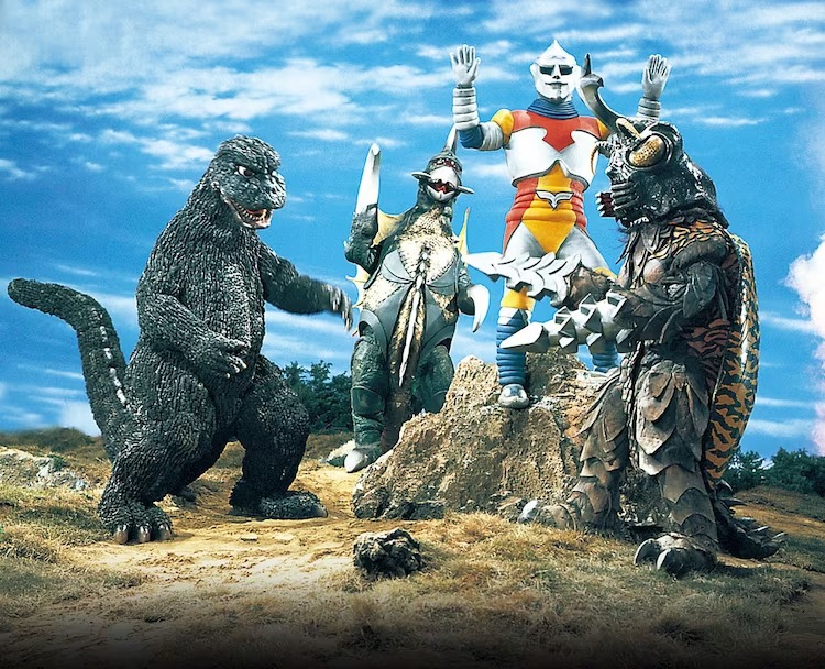 A promotional set photo for the 1973 film, Godzilla vs. Megalon, featuring the suit actors in costume for Godzilla, Gigan, Jet Jaguar, and Megalon all posing pugnatiously on a set made up to look like a rural battlefield in front of a blue sky laced with status clouds.