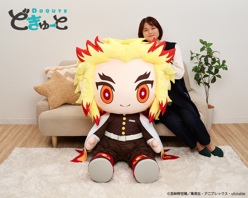 A promotional image for the DoQute 2XL Plush Toy of Kyojuro Rengoku from Demon Slayer: Kimetsu no Yaiba by Taito Corporation. The image depicts the jumbo-sized plush toy next to a Japanese model seated on a sofa for a size comparison.