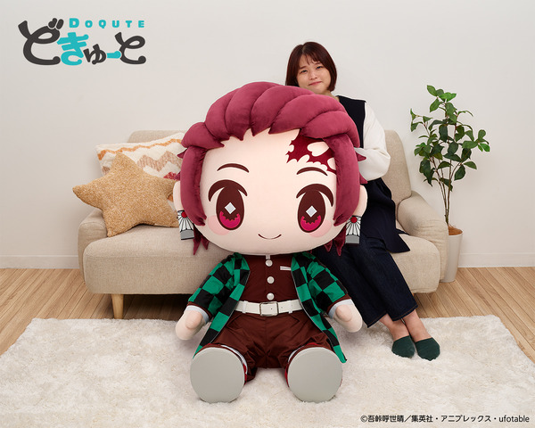 A promotional image for the DoQute 2XL Plush Toy of Tanjiro Kamado from Demon Slayer: Kimetsu no Yaiba by Taito Corporation. The image depicts the jumbo-sized plush toy next to a Japanese model seated on a sofa for a size comparison.