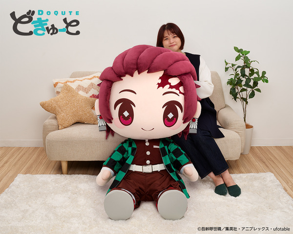 Ein Werbebild für das DoQute 2XL Plüschtier von Tanjiro Kamado aus Demon Slayer: Kimetsu no Yaiba von Taito Corporation.  Das Bild zeigt das riesige Plüschtier neben einem japanischen Model, das zum Größenvergleich auf einem Sofa sitzt.