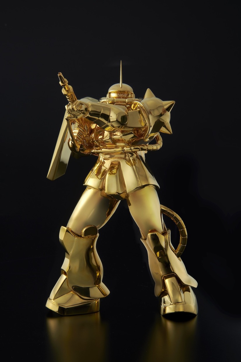 A promotional image of the U-Works Mobile Suit Gundam Solid Gold Statue Char's Custom Zaku II, featuring the solid gold mecha posed hoisting its rifle in a combative manner, indicating the pilot has just fired off a round.