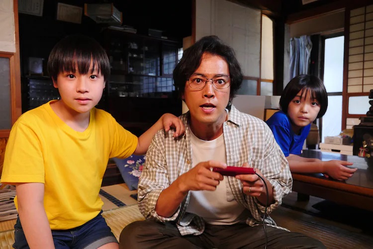 The characters of Kenichi Nakaoka, Ryousuke, and Yuuki gather around a haunted copy of Dragon Quest II: The Luminaries of Legend in a scene from the upcoming "Scroll of Restoration" episode of the Tales of the Unusual TV drama.