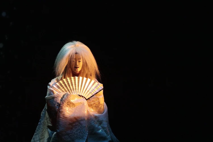A promotional photo of a masked performer from the VR Noh Ghost in the Shell stage play. The performer is dressed in a flowing white kimono, a traditional Noh mask, and a white wig while carrying a fan, and the stage is dark except for the spot light shining upon the actor.