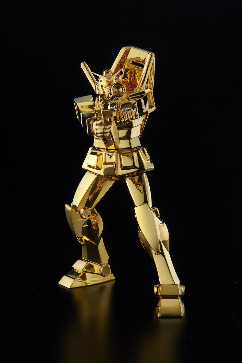 Una imagen promocional del U-Works Mobile Suit Gundam Solid Gold Statue RX-78-2 Gundam (Beam Rifle Ver.), Con el mecha de oro macizo levantando su rifle de rayos en una postura que indica que el piloto está listo para disparar.