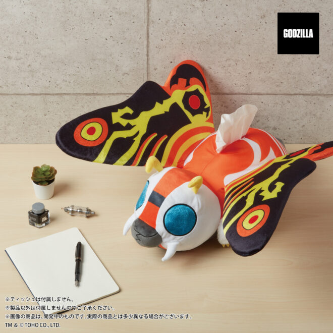 A promotional image of the Mothra (Imago) Tissue Case product from Premium Bandai featuring a close-up view of the product viewed from above while it is placed on a desk next to a potted plant, and inkewell, some stationery, a pen, and a tiny model of the Oxygen Destroyer from the original 1954 Godzilla film.