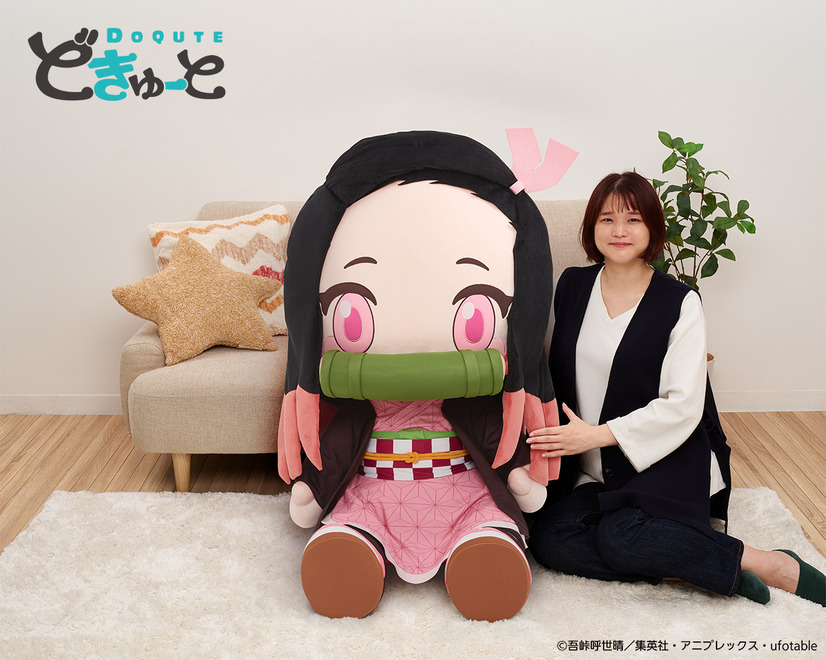 A promotional image for the DoQute 2XL Plush Toy of Nezuko Kamado from Demon Slayer: Kimetsu no Yaiba by Taito Corporation. The image depicts the jumbo-sized plush toy next to a Japanese model seated in front of a sofa for a size comparison.