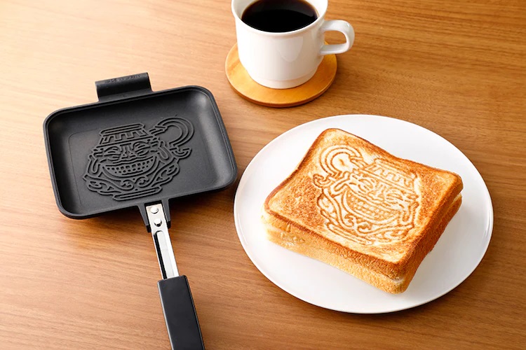 Ein Werbebild für den Pot of Greed Hot Sandwich Maker mit dem namensgebenden Küchenwerkzeug, einer Tasse Kaffee und einem Teller mit einem gegrillten Käsesandwich darauf.