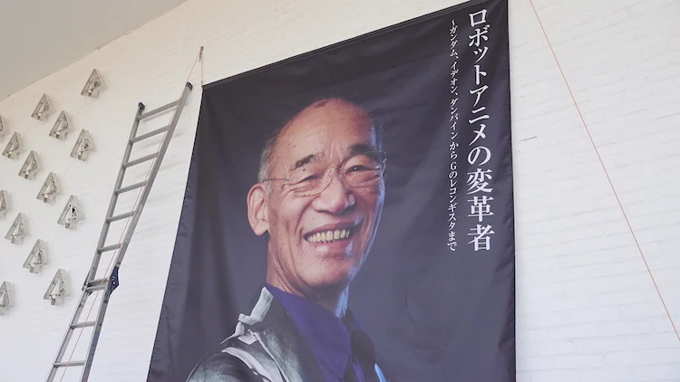 A promotional photo from the Yoshiyuki Tomino's World art exhibition, featuring a large banner of Tomino flashing a warm smile.