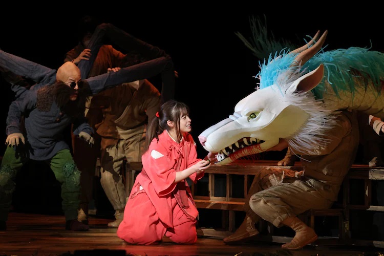 A promotional photo from the stage play adaptation of Spirited Away depicting actor Kanna Hashimoto as Sen interacting with the characters of Kamaji and Haku as depicted by actors in make-up and puppets operated by black-clothed puppeteers. 