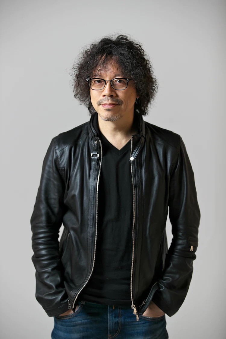 A promotional photograph of manga author Naoki Urasawa. Urasawa wears a leather jacket, a black T-shirt, and jeans. He smiles casually with his hands in his pockets.