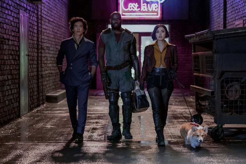 A promotional image for Netflix's upcoming Cowboy Bebop live-action series, featuring the main cast of John Cho, Musafa Shakir, and Daniella Pineda in full costume and make-up as Spike Spiegel, Jet Black, and Faye Valentine, respectively. The three bounty hunters stride down a darkened alley while walking with Ein, a super intelligent Welshi corgi dog, on a leash.