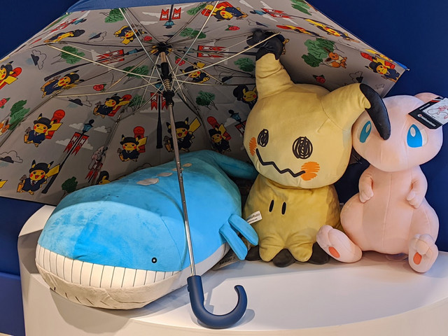 A Pokémon Center London umbrella next to the deluxe plushies of Mimikyu, Mew and Wailord