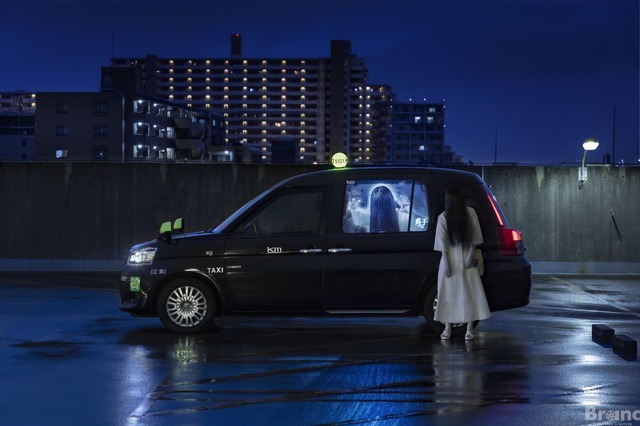 A promotional image from the upcoming "Sadako Taxi" collaboration featuring the ghostly Sadako posing next to a Sadako-themed taxi cab in Tokyo at night.