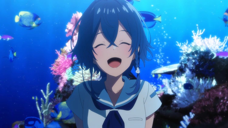 Heroine Kukuru Misakino smiles in front of an aquarium filled with coral and tropical fish in a scene from the upcoming Aquatope of White Sand TV anime.