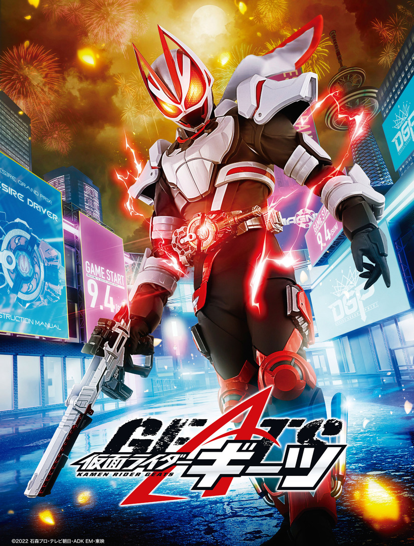A promotional image for the upcoming Kamen Rider Geats live-action tokusatsu TV series, featuring the titular Kamen Rider Geats, a masked superhero with a white fox motif, standing in brightly lit street of a futuristic city-scape while fireworks explode in the night sky above him.