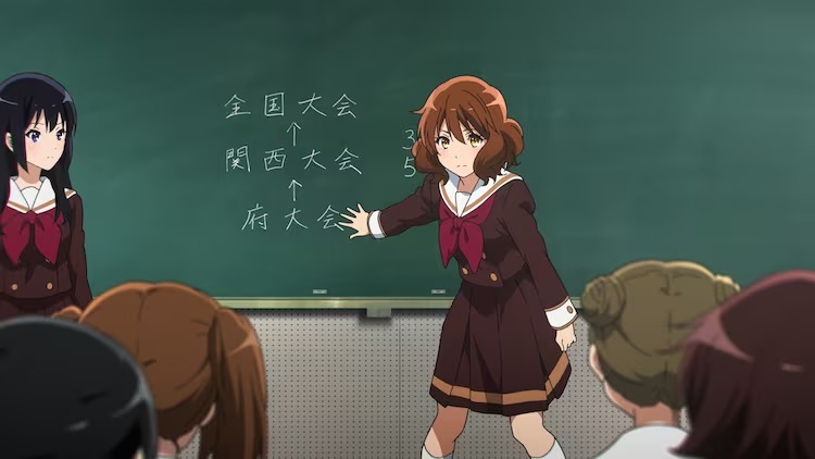 Kumiko Oumae gestures at the blackboard while addressing the brass band club in a scene from the upcoming Sound! Euphonium theatrical OAV.