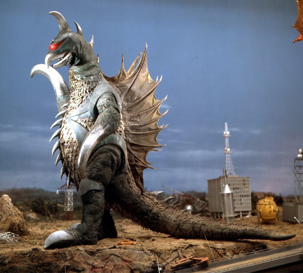 Ein Werbebild des riesigen Monsterschurken Gigan aus dem Set des Films Godzilla vs. Gigan von 1972.  Gigan ist ein kybernetisches Monster mit Haken als Arme, einem schnabelförmigen Kopf, der mit Stacheln geschmückt ist, einem einzigen leuchtend roten Auge und einer Kreissäge in seinem Bauch.