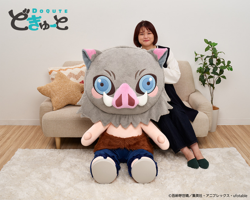 A promotional image for the DoQute 2XL Plush Toy of Inosuke Hashibira from Demon Slayer: Kimetsu no Yaiba by Taito Corporation. The image depicts the jumbo-sized plush toy next to a Japanese model seated on a sofa for a size comparison.