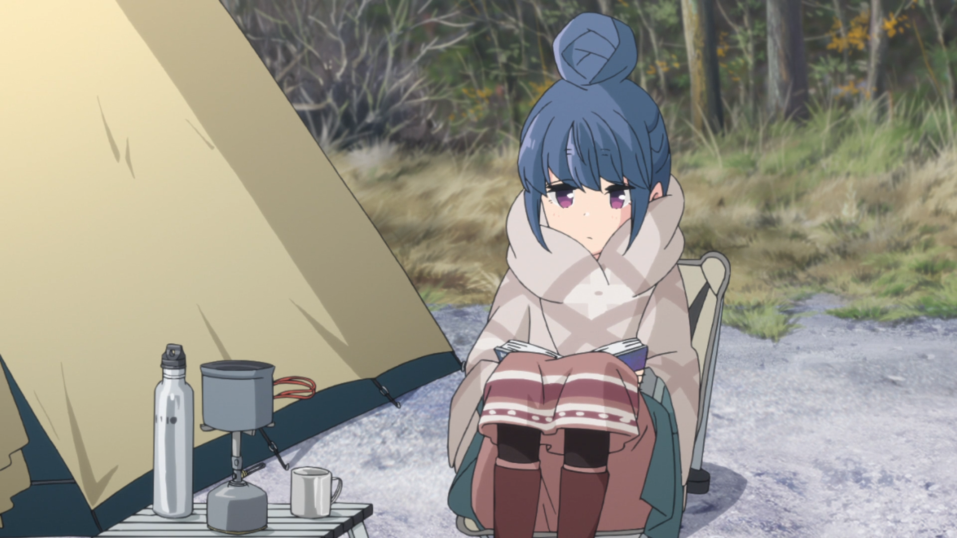 Rin Shima se acurruca con un libro y un paquete de calefacción de bolsillo mientras se relaja en su silla portátil en su campamento en una escena del anime de televisión Laid-Back Camp.