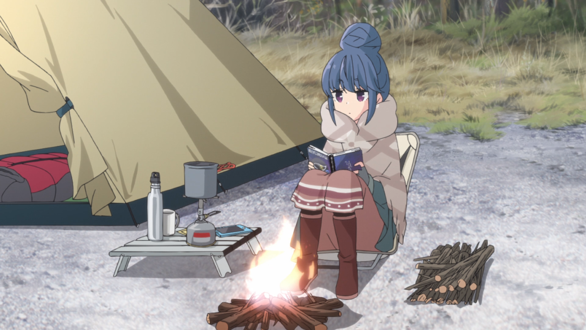 Surrounded by her tent and modern camping gear, Rin Shima enjoys some solo camping by reading a book near a campfire during a cold winter day in a scene from the 2018 Laid-Back Camp TV anime.