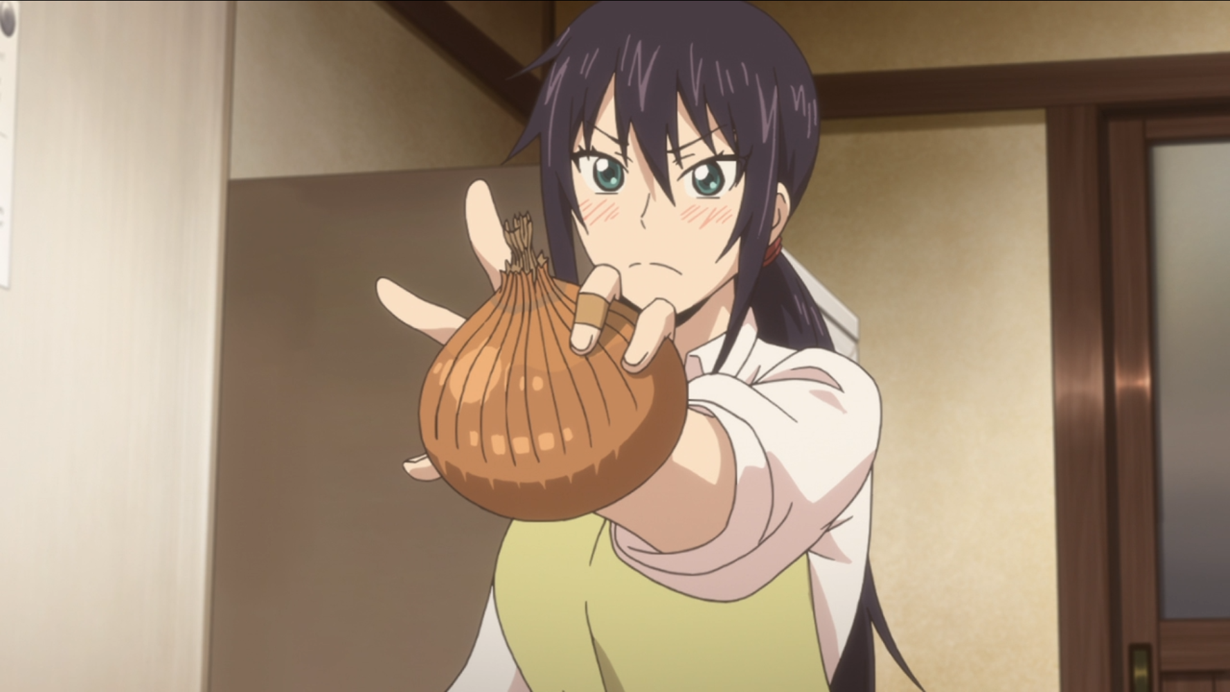 Kotori Iida schwenkt mit ihren verbundenen Fingern, die auf ihre früheren erfolglosen Kochversuche hinweisen, eine große rohe Zwiebel in einer Szene aus dem TV-Anime Sweetness & Lightning 2016.