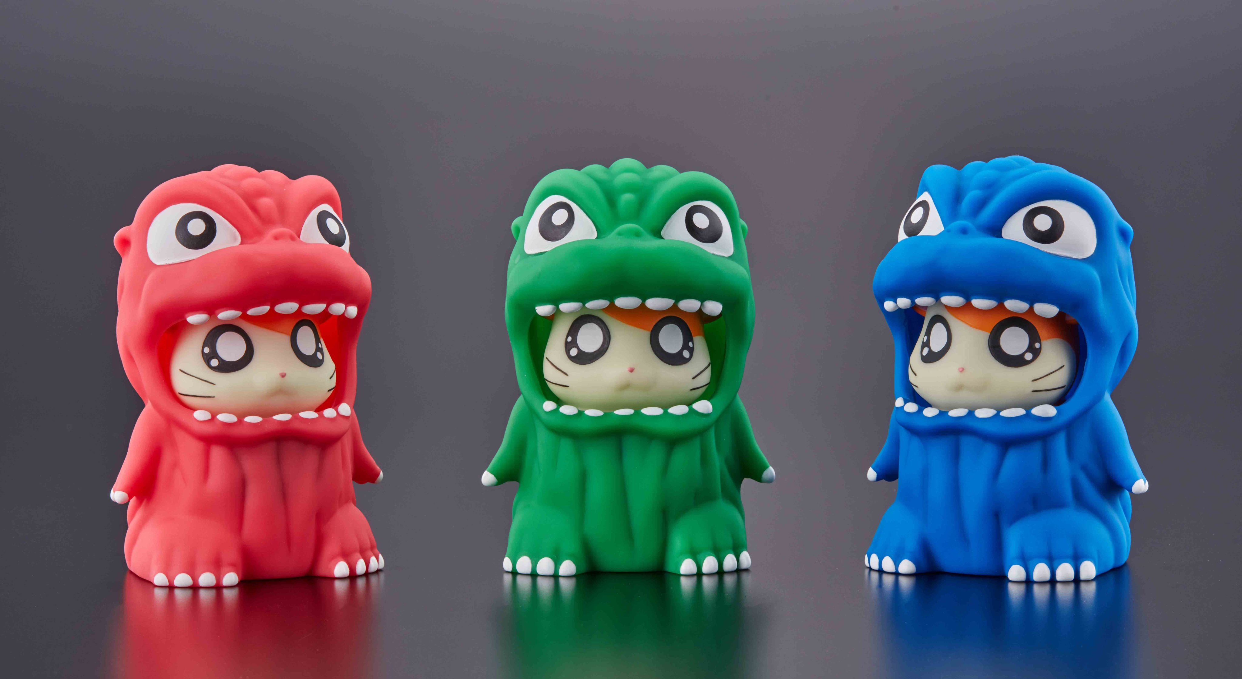 A promotional image featuring three color variants (red, green, and blue) of the Godzihamkun collaboration character, featuring Hamtaro the hamster wearing a cute Godzilla costume.